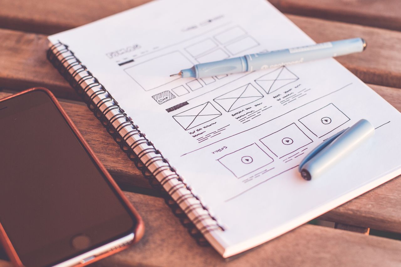 Gros plan sur un carnet avec des esquisses de wireframe et un smartphone posé sur un bureau en bois.